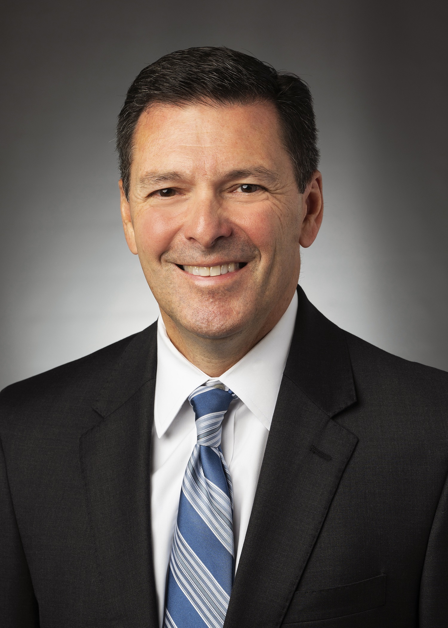 Smiling man in a dark suit with a blue striped tie, studio portrait.